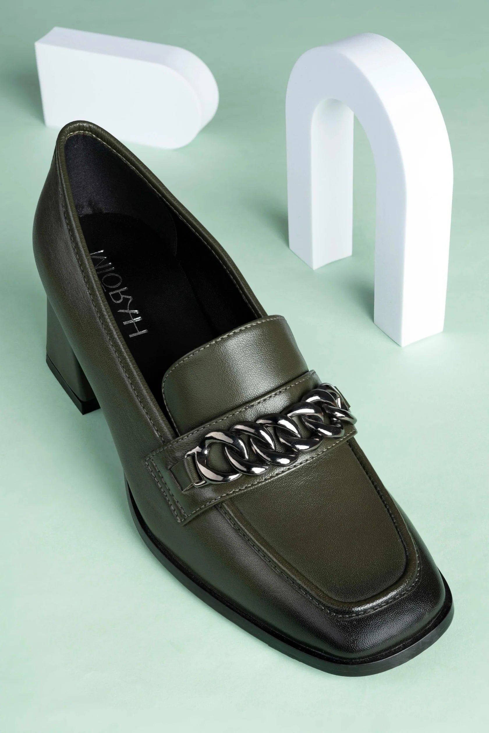 Dark brown loafer shoe with chain detail on a light green background