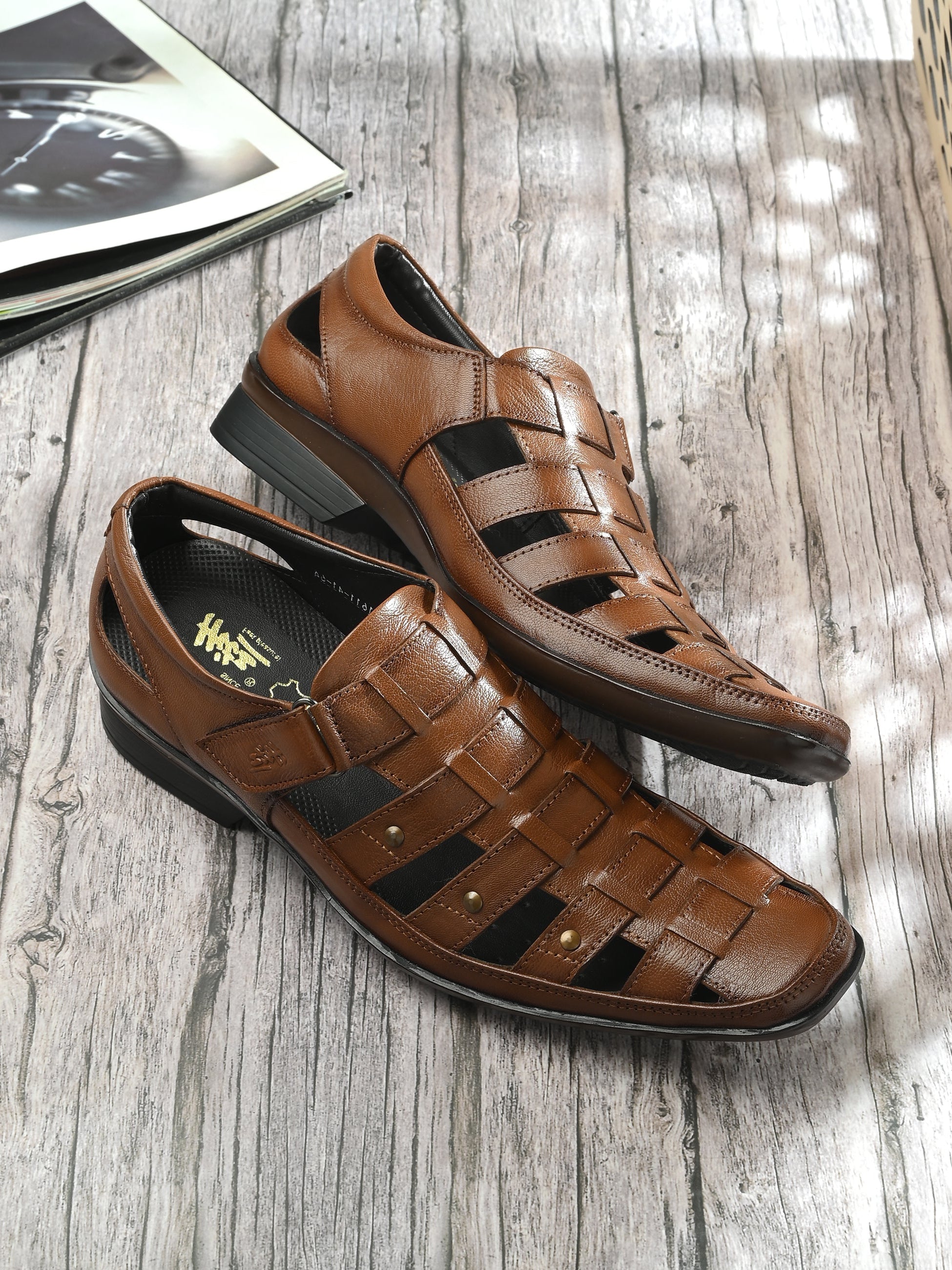 A tan brown leather Roman sandal with a closed toe and strap details.